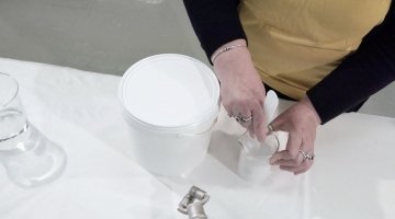 A pair of hands reach over a table, using a small white spatula to stir paste in a beaker. A large white bucket rests on the table beside the beaker. 