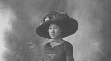 A black and white image of a Chinese woman in a more traditionally European style of dress, also wearing a hat and carrying a satchel 