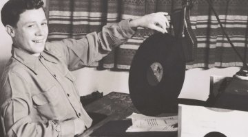 At the left of the photo, a young man with short dark hair is sitting down at a radio desk, smiling out at the camera and with his left hand he balances a record on its edge on the desk.