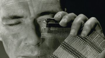 A man's face from forehead to shoulders fills the image. He's holding up a rectangular piece of woven metal strips to his left eye to inspect them