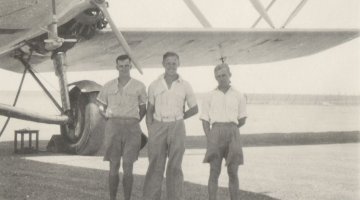 Three men stand in the centre of the sepia photograph, with the nose and left wing of a biplane visible behind them
