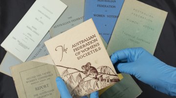 Two hands in blue latex gloves hold up a small booklet with the title 'The Australian Federation of Womens Societies'. Other booklets relating to the Australian Federation of Women Voters lie flat on the table underneath.