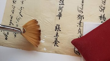 A conservator's fan brush hovers over delicate paper showing Chinese characters, with a red pillow weight on the righthand side of the paper.