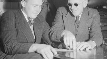 Two men sit side by side. The man on the right is teaching the other man to read in braille by guiding his hand along the words. The teacher wears small round sunglasses. Both men are wearing pinstripe suits and smiling.