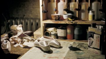 Ballet slippers are sitting on newspaper on a table with paints and other craft items on a shelf behind.