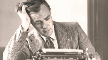 A black and white photo of Stewart Cockburn with his head in his hand, sitting at a typewriter