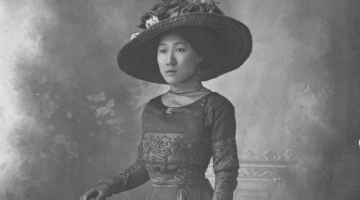 A black and white photograph of a Chinese Australian woman dressed in an 1800s dress and hat