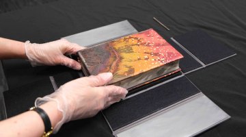 A painted hardback book is being placed into a black box by a pair of gloved hands