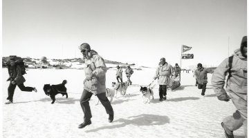 A group of people and dogs in thick snow gear run through the snow