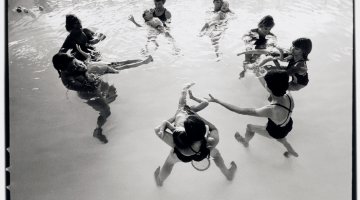 A black and white landscape of women floating in a pool in a circle with toddlers accompanying them. 