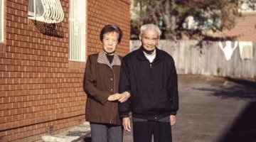 An elderly couple stand arm in arm outside a brick building