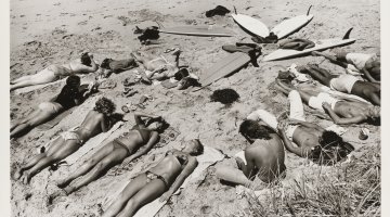 A black and white landscape of a group of sunbathing people lying on a beach with multiple surfboards lying on the ground together. 
