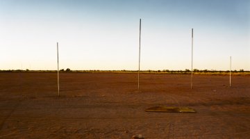 Football goal posts on a red dirt field with a clear blue sky, a single football book abandoned in the front.
