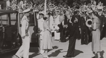 A black and white image of Queen Elizabeth and Prince Philip getting out of a car and shaking hands with officials. There are many military personnel in the background.