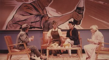 Four people sitting on couches and armchairs around a coffee table. There is a large artwork on the wall behind them of a man in a hat holding a bottle of softdrink.