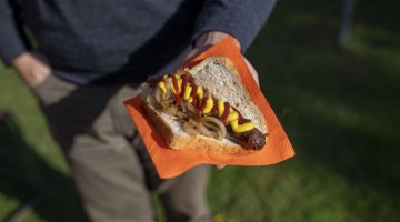 The image of a person from the waist down. In one hand held out in front of them they are holding an orange paper napkin with a sausage sandwich on it.