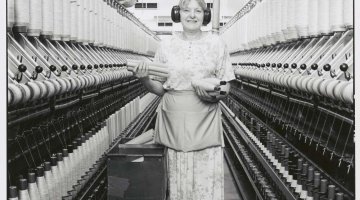 A woman holding some large spools of thread, standing in between two long factory machines for thread. Black and white.