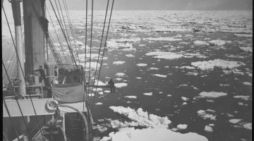 A black and white photograph. The mast and bow of a ship are on the left hand side of the image and water littered with ice chunks are on the right. This image is taken from an elevated position. 