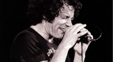 A black and white image of a man holding a microphone on a stand with 2 hands. He is holding the mic close to his mouth and has his eyes shut. He is wearing a black t-shirt and jeans.