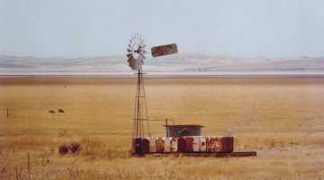 A windmill stands in a paddock surrounded by dry, yellow grass. Lake George in New South Wales is visible in the background.