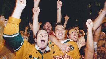 a crowd of Australian fans wearing green and gold jerseys cheer on, hands raised and shouting. It's nighttime and the photographer has captured some of the motion blur enhancing the evening's excitement.