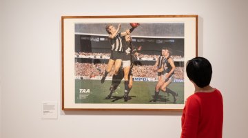 A person in a red jumper standing in front of a framed image that shows AFL players mid-game.