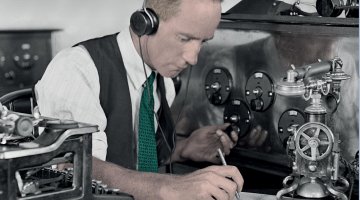 A man wearing old fashioned headphones is sitting at a table. He is writing on a notepad and is surrounded by lots of old fashioned communications equipment.