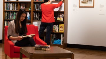 Woman sitting on a red armchair reading in front of large bookshelf with another woman looking at a book placed on the shelf beside a framed image of Shane Warne