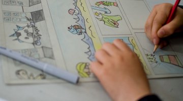 A close-up of a comic strip drawing, with two hands. One hand is holding a pencil and the other is holding the page.