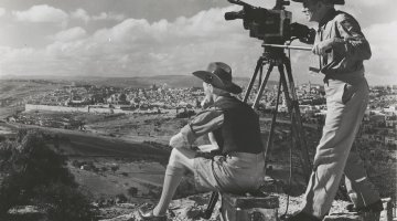 A black and white image of two men on a hilltop overlooking a valley. Both men are wearing hats. One man is sitting on a box. The other is standing up behind an old 1940s style film camera that is on a tripod.
