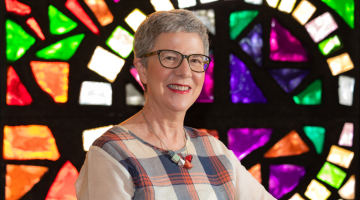 Woman with short grey hair and black glasses wearing checkered white, blue and red shirt, smiling and standing in front of colourful stained glass windows 
