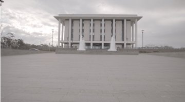 Exterior of tall building with grey columns and a fountain out the front, on a cloudy day. 