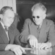 Two men sit side by side. The man on the right is teaching the other man to read in braille by guiding his hand along the words. The teacher wears small round sunglasses. Both men are wearing pinstripe suits and smiling.