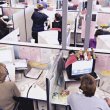 Rows of desks in a call centre. Employees are working at a number of the desks. 
