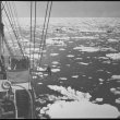 A black and white photograph. The mast and bow of a ship are on the left hand side of the image and water littered with ice chunks are on the right. This image is taken from an elevated position. 