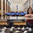National Library of Australia 2022 scholarship recipient, Madeleine Pugin, stands smiling behind a glass cabinet containing Indigenous aluminium cast artefacts.