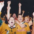 a crowd of Australian fans wearing green and gold jerseys cheer on, hands raised and shouting. It's nighttime and the photographer has captured some of the motion blur enhancing the evening's excitement.