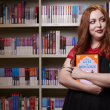 Woman with red hair standing in front of a bookshelf of romance books, holding a tablet with the cover of a romance book on the screen. She is facing forward with her back to the bookshelf, looking to the side and slightly smiling
