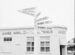 black and white photograph shows a white wooden signpost with arms pointing to Honolulu, Calcutta, Nadi, Sydney, Auckland and the shell service station, in front of a white building with Lane's Agricultural Chemicals painted on the side.