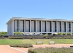 A view of the exterior of the National Library of Australia building. Green grass and red dirt is visible in the foreground.