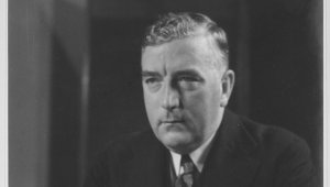 Australian Prime Minister Robert Menzies sits at a desk holding several pieces of paper. He is looking melancholic and stares off camera to the left. He is a middle-aged white man with blonde hair swept to one side. He wears a dark suit and tie with a dark tie. There is a black Bakelite telephone on the table in front of him