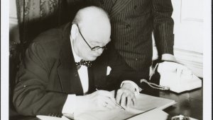 British Prime Minister Winston Churchill is seated at a table wearing glasses. He is signing a document. Standing behind him and leaning over his shoulder is Australian Prime Minister Robert Menzies. Both men are wear dark suits with white shirts. Menzies is wearing a dark tie, Churchill a bow tie.
