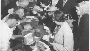 A black and white photograph showing a group of women shopping for food and supplies using wartime coupons. There are three shopkeepers behind the counter handing out goods and taking coupons