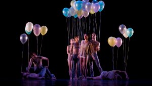 A troupe of dancers illuminated on a blacked out stage. They are holding colourful balloons. Some actors are laying on the floor, most are standing.