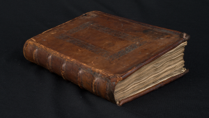 A closed book. It is sitting on a black velvet cloth. The book is bound in brown leather. The front cover and spine are carved and moulded. The front cover is very scratched and worn.