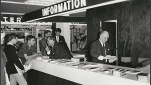 A black and white photograph of a group of men standing at a white information desk. Above the desk is a large sign that says INFORMATION. Men behind the counter and speaking and hanging out brochures.