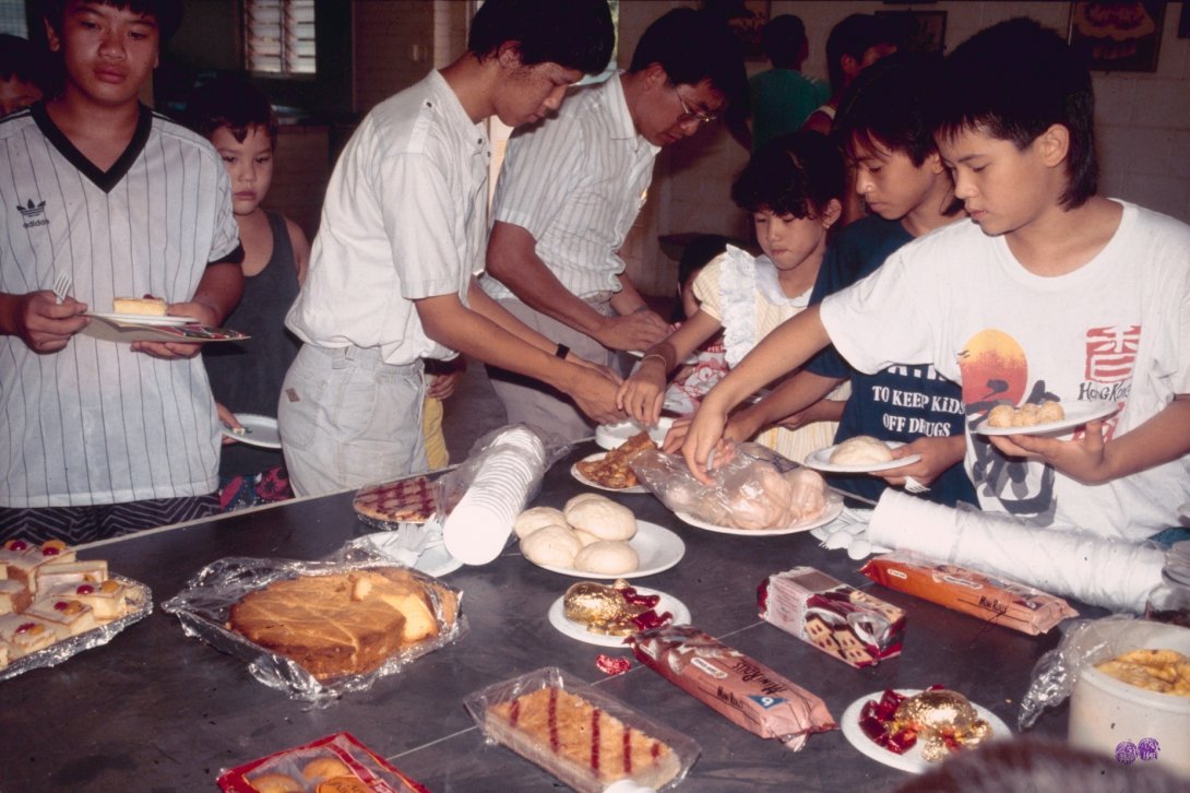 A group of 7 people ranging from children to adults are gathered around the far side of a table in the foreground. The table is laden with desserts.
