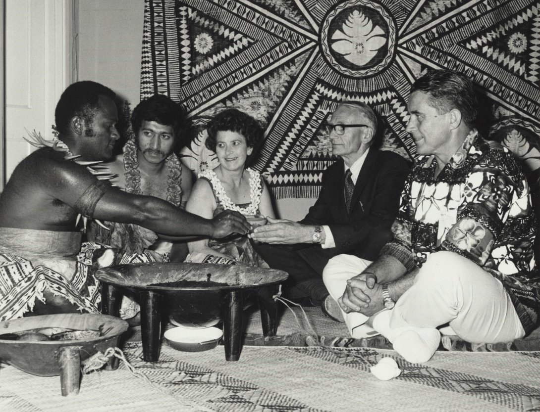 Five people sit cross legged on the floor around a small cooking basin on legs. One white woman sits in the middle, with two Fijian men in traditional dress on the left and two white men on the right.