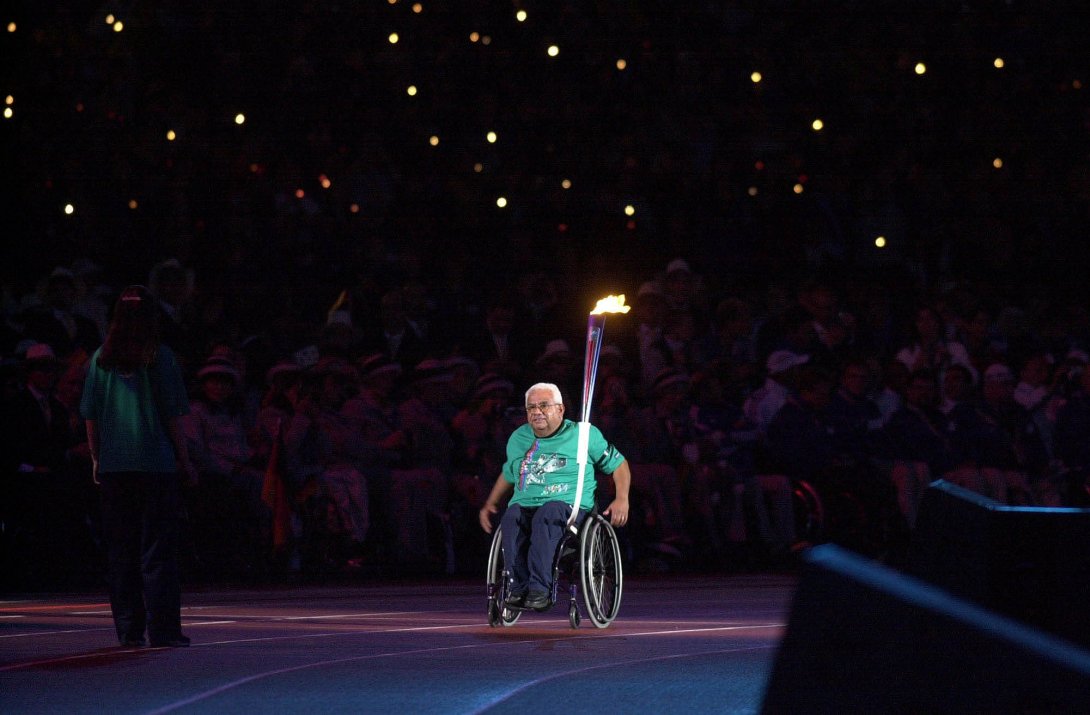 At the centre of an image a man seated in a wheelchair, wearing dark blue trousers and a green t-shirt, is wheeling himself along an athletics track. An Olympic torch is attached to his chair on the right. It is nighttime, and while a spotlight shines on the man, a crowd behind him in stadium seating is mostly in the dark, with a few lights visible, as if from candles.