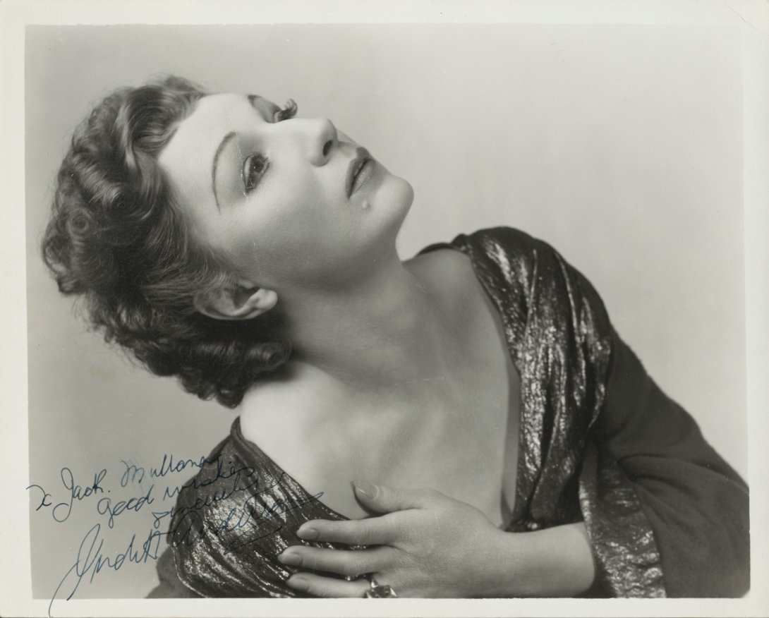 A woman leans her head back, looking up out of the photograph. She has short, wavy hair and is wearing a long-sleeved, shimmery dress with a low v-neckline. 